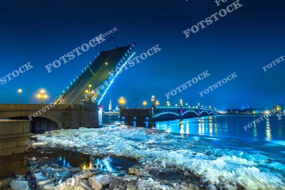 На фотографии изображён разведённый Троицкий мост, освещённый ночной подсветкой. Льдины на поверхности Невы подчёркивают зимний характер города. Вдалеке видны огни Петропавловской крепости, создающие атмосферу исторического Санкт-Петербурга. Троицкий мост — один из символов города, соединяющий его берега и украшающий зимний пейзаж.